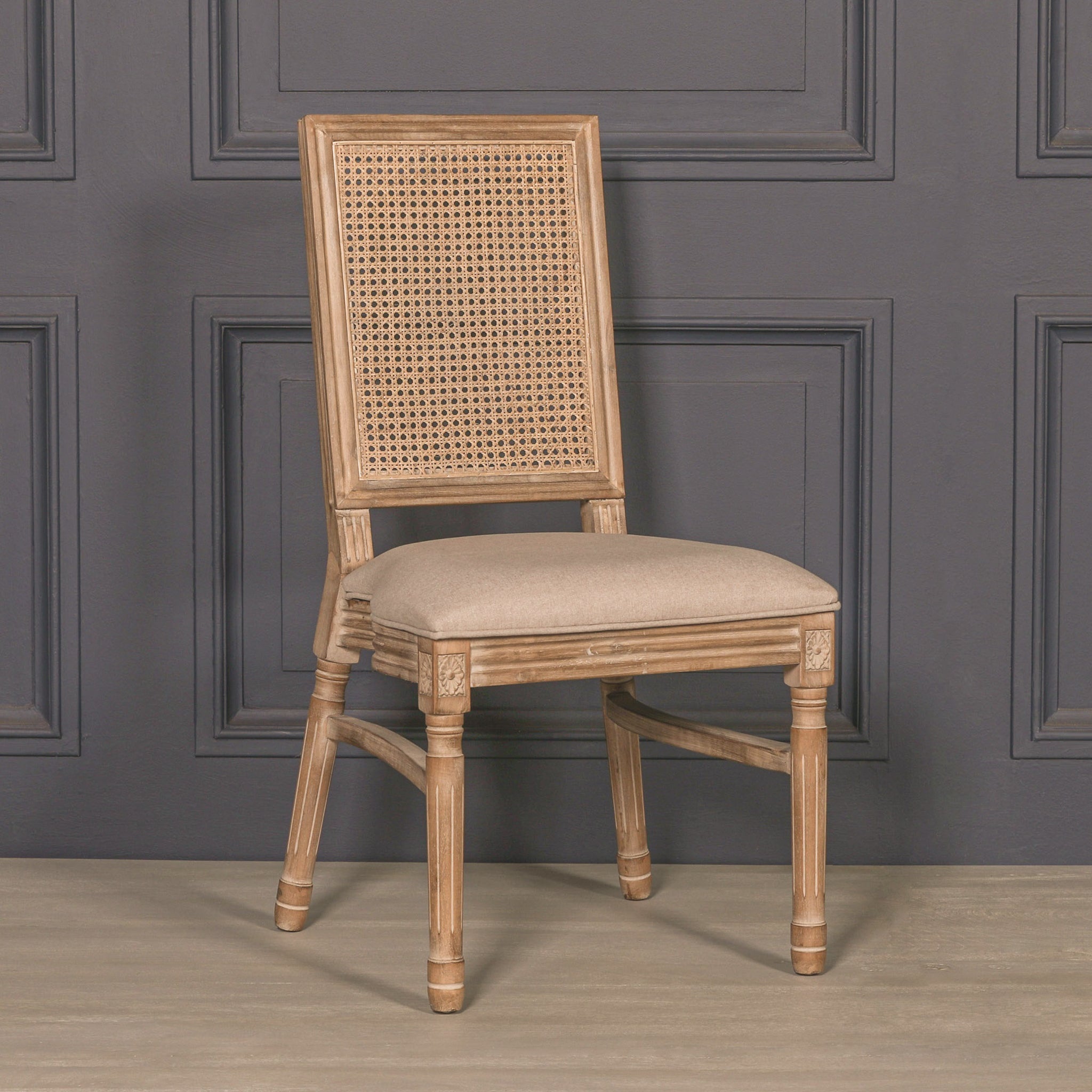 Three-quarter view of the Camille French Louis Rattan Back Dining Chair with woven rattan back, linen seat cushion and carved wooden frame against a charcoal panelled wall.