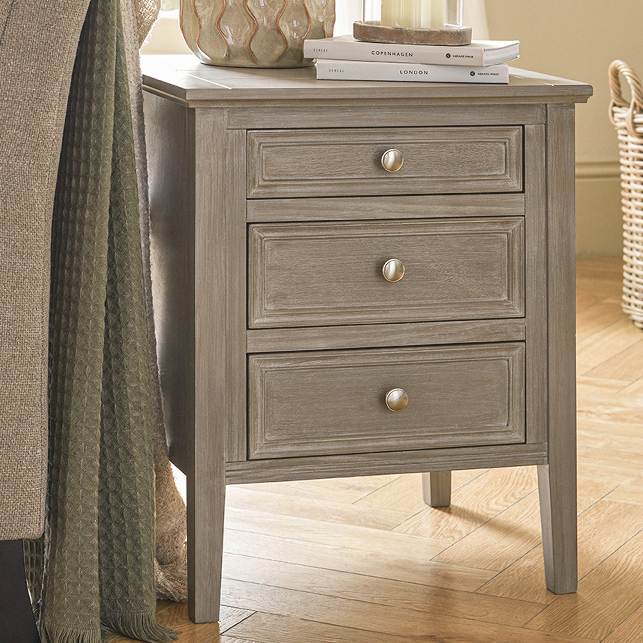 Camille grey wooden bedside table with three drawers styled beside a bed, featuring a painted wood finish, brushed metal knobs and spacious bedroom storage, perfect as a bedside cabinet, nightstand or bedside chest of drawers.