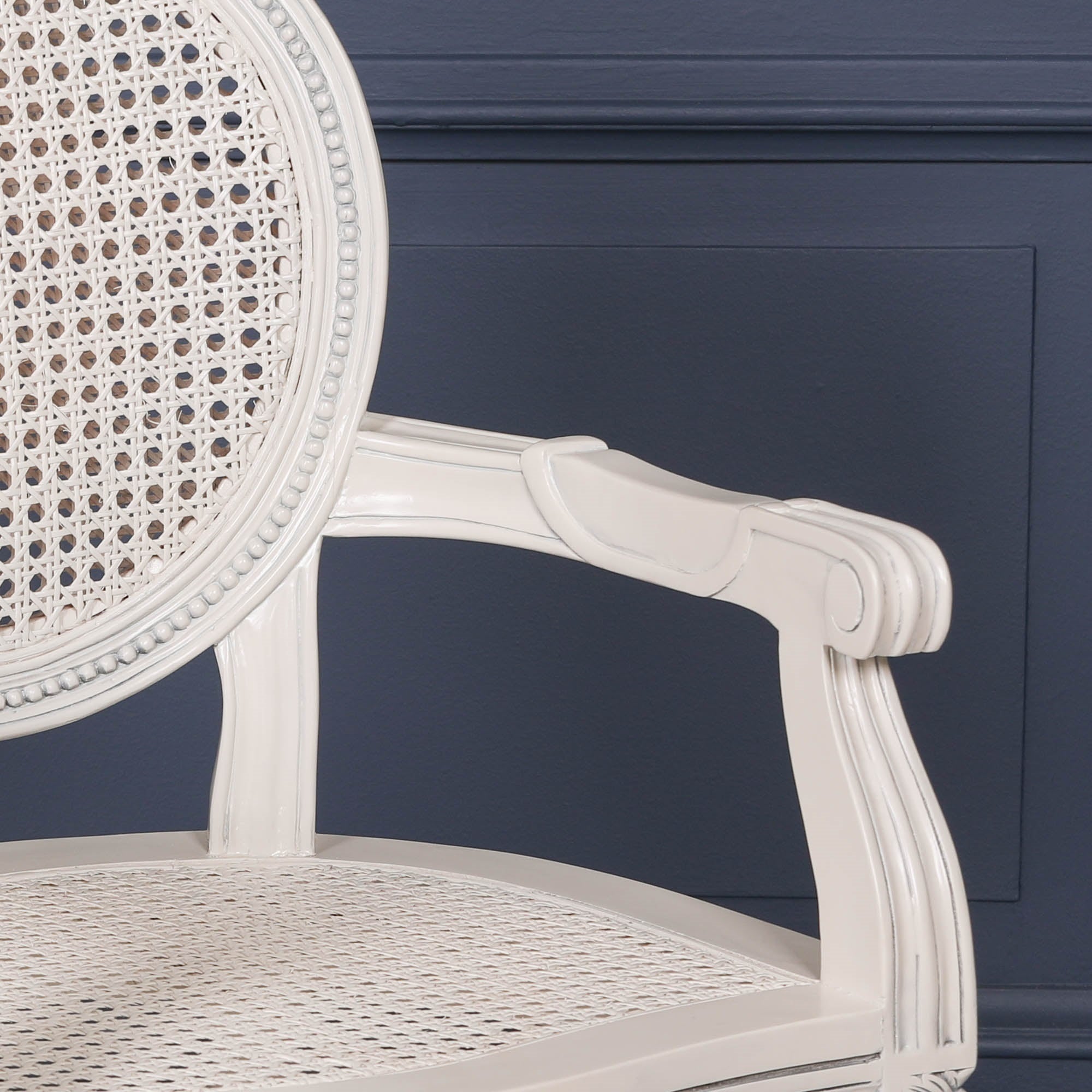 Close-up of the Céleste French Chateau Rattan Dining Arm Chair in Off-White Mahogany featuring the carved scroll armrest, woven rattan seat, oval cane backrest and distressed painted mahogany finish.