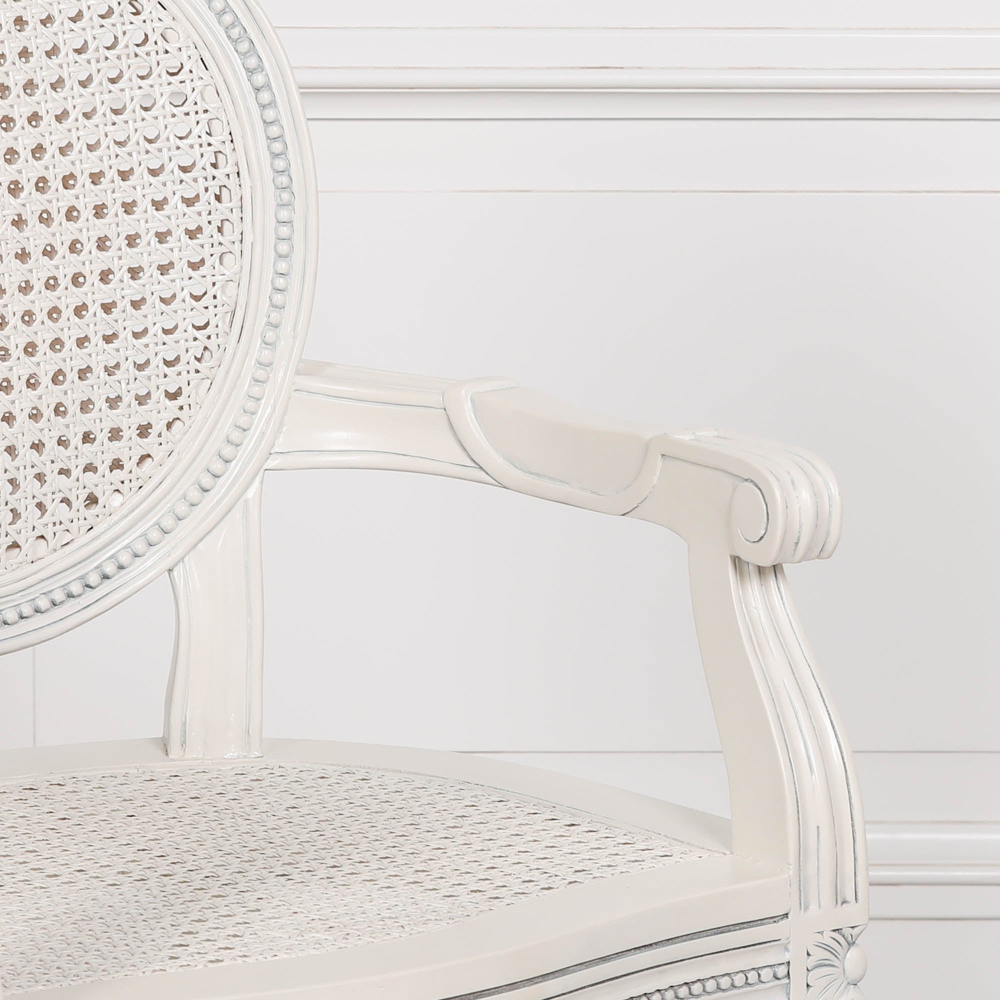 Close-up of the Céleste French Chateau Rattan Dining Arm Chair featuring the carved scroll arm, woven rattan seat, oval cane backrest and distressed off-white mahogany finish.