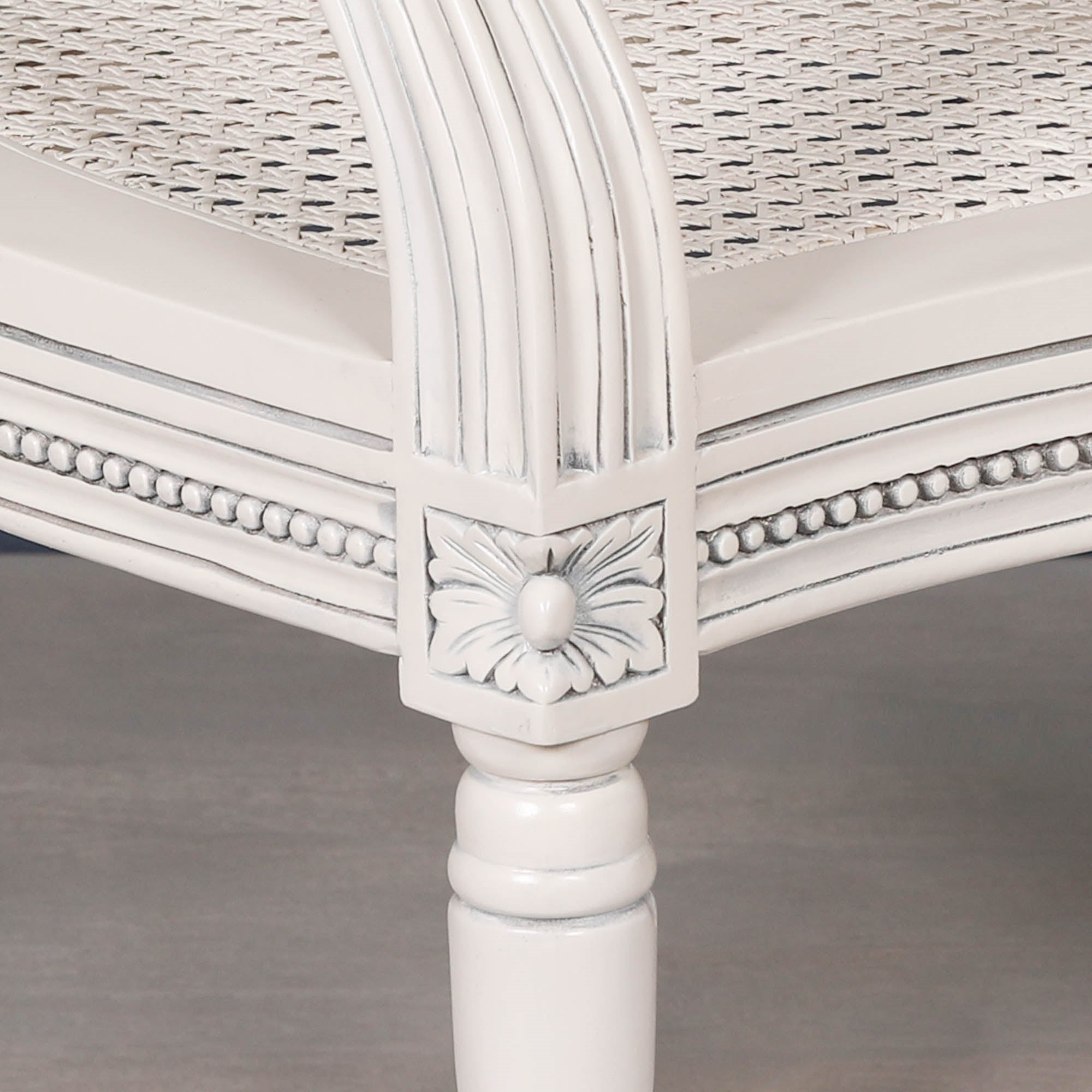 Detailed close-up of the Céleste French Chateau Rattan Dining Arm Chair highlighting the hand-carved floral corner block, beaded trim, turned mahogany leg and woven rattan seat in an off-white finish.