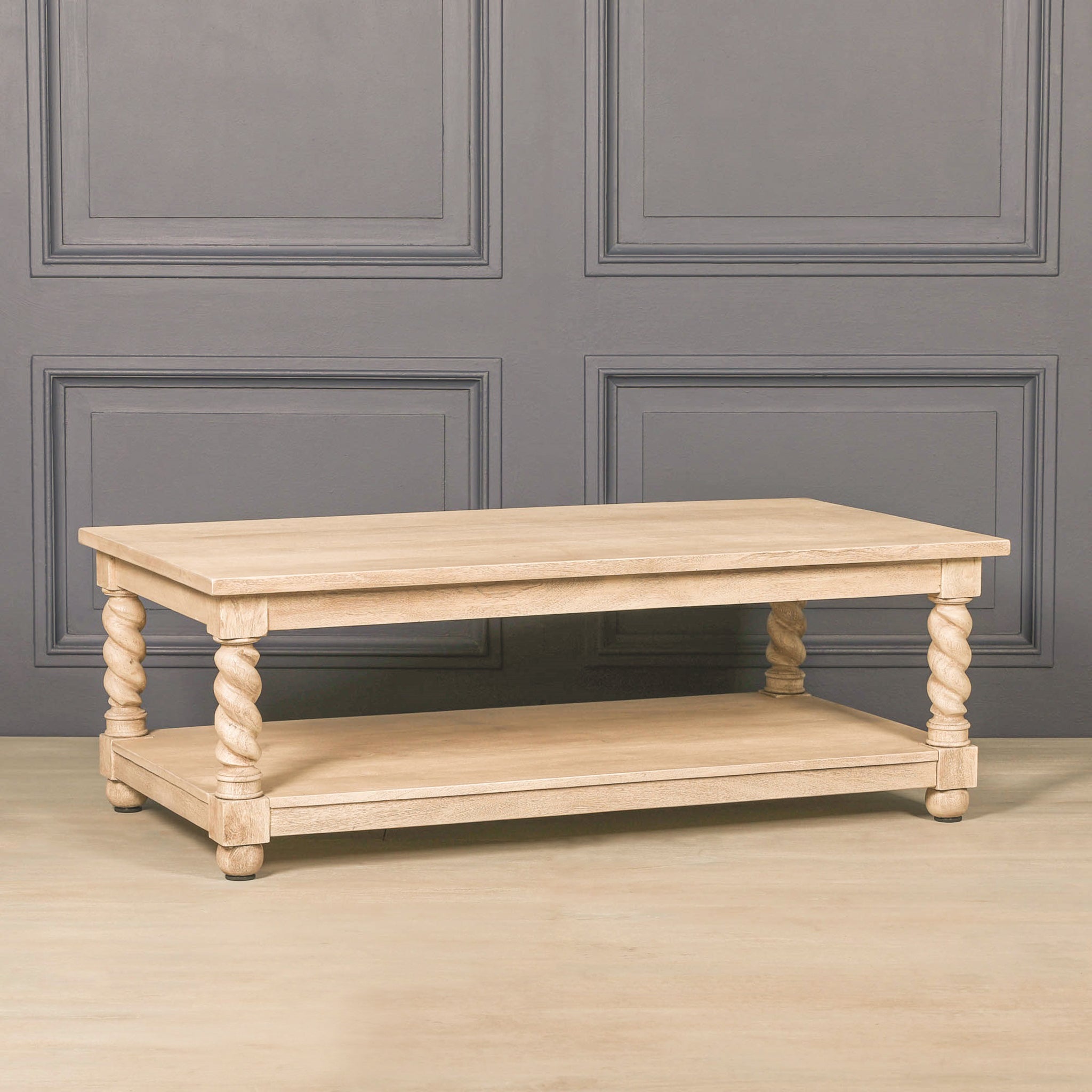 Front view of the Éloi sculptural wood coffee table against a dark panelled wall, highlighting its rectangular top, four decorative twisted legs, lower shelf and softly washed acacia wood finish.