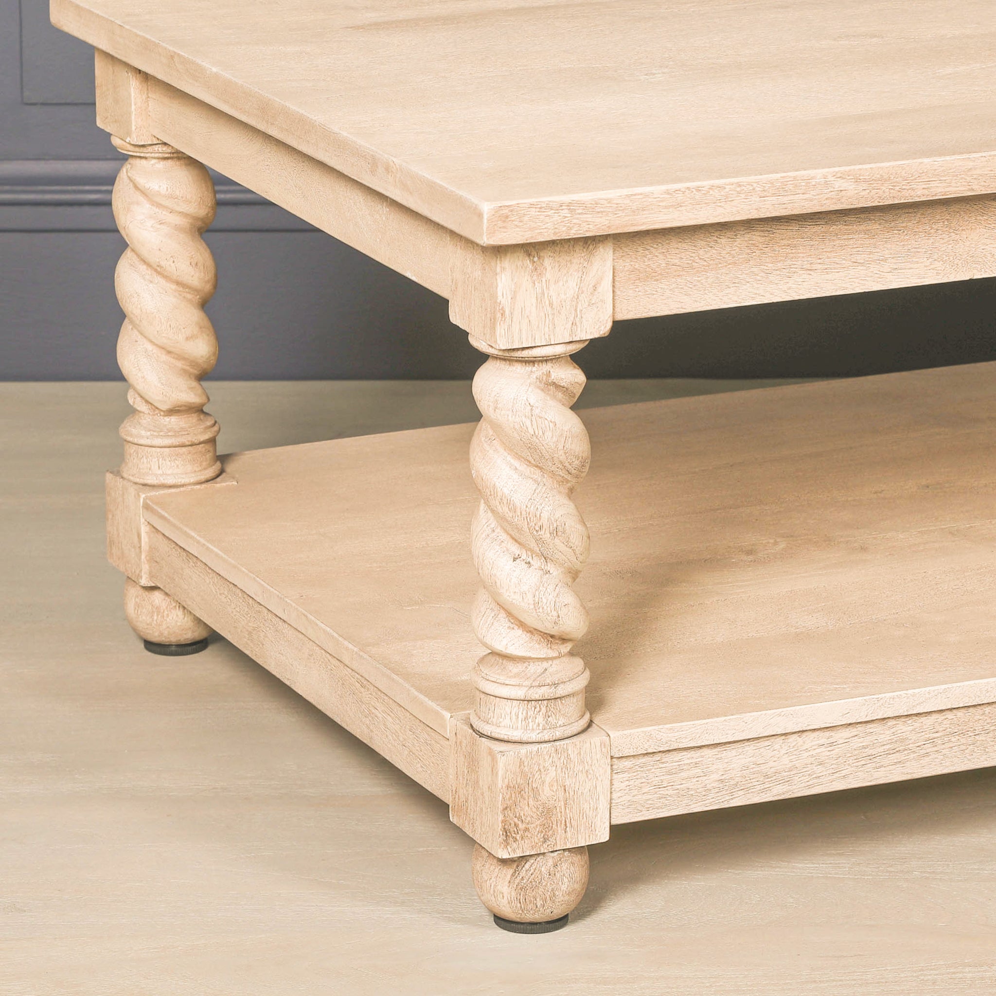 Close-up of the Éloi solid acacia wood coffee table showing the sculptural hand-carved twirl legs, rectangular lower shelf, rounded feet and natural grain of the light washed wood finish.