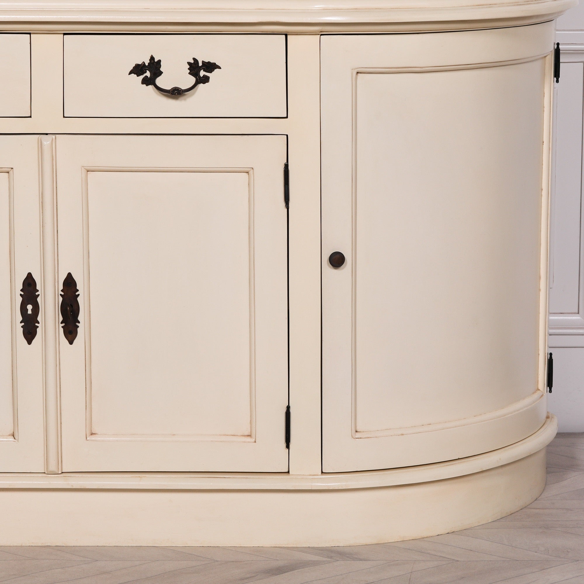 Close-up of Geneviève aged cream mahogany cabinet curved storage base against a white panelled wall.