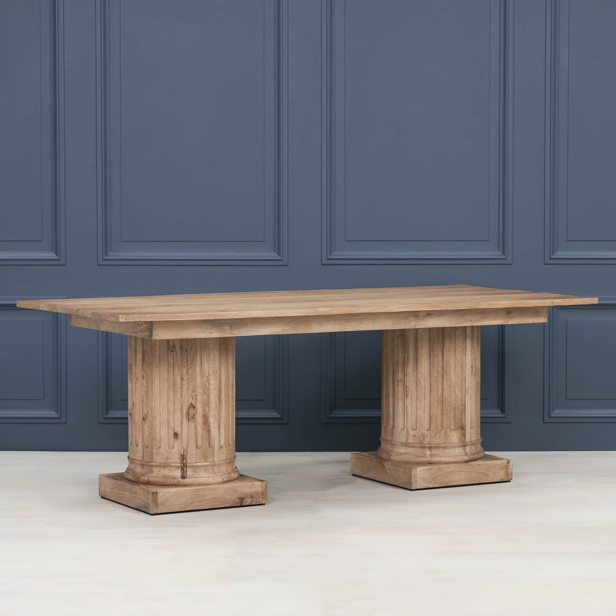 Angled view of the Henri 220cm Rustic Mango Wood Rectangular Column Dining Table against a blue panelled wall, highlighting the rustic solid mango wood construction, rectangular tabletop and architectural twin column pedestal bases that seat 8 to 10 people.
