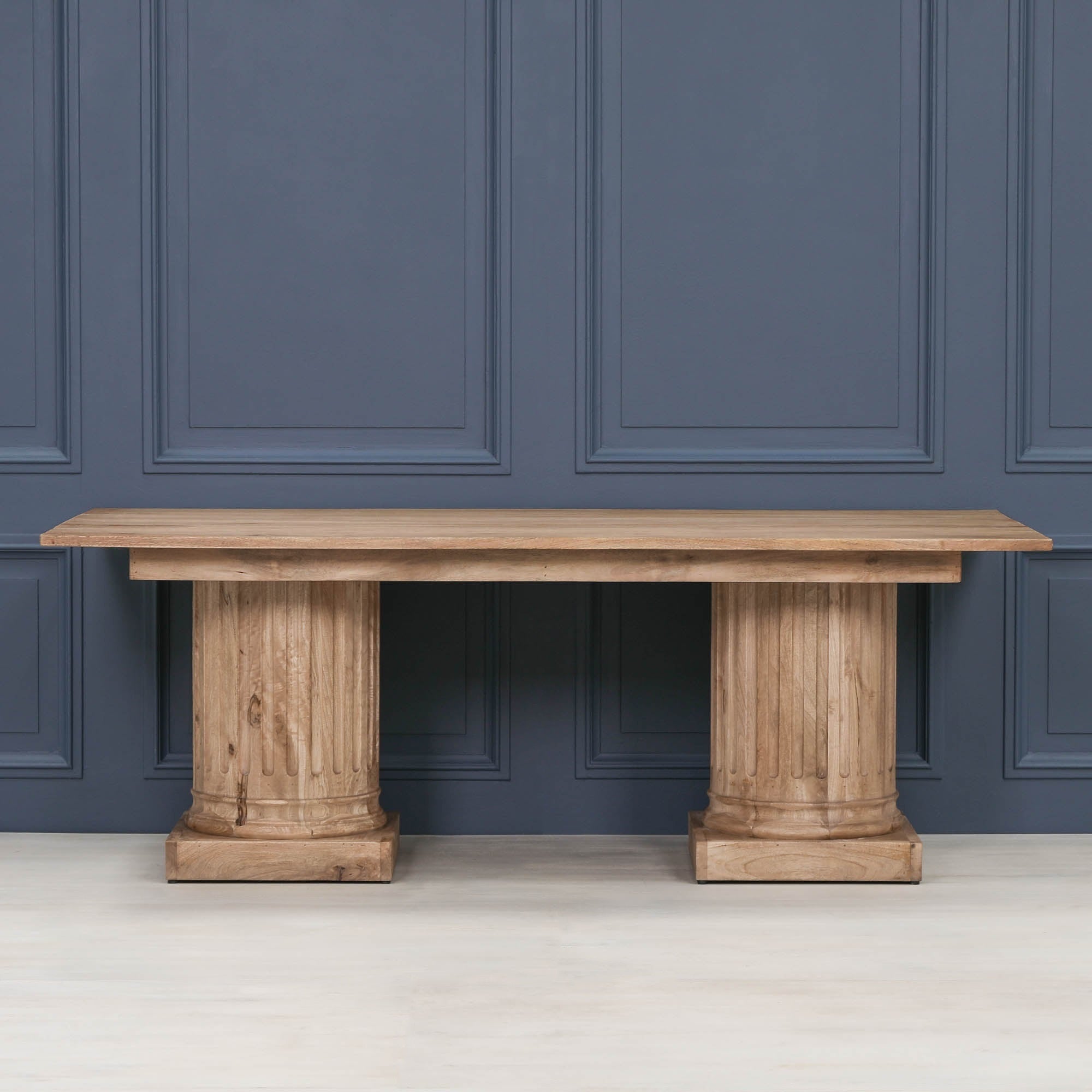 Front view of the Henri 220cm Rustic Mango Wood Rectangular Column Dining Table against a blue panelled wall, showcasing the rustic mango wood finish, fluted column pedestal bases, generous rectangular tabletop and timeless contemporary farmhouse design.
