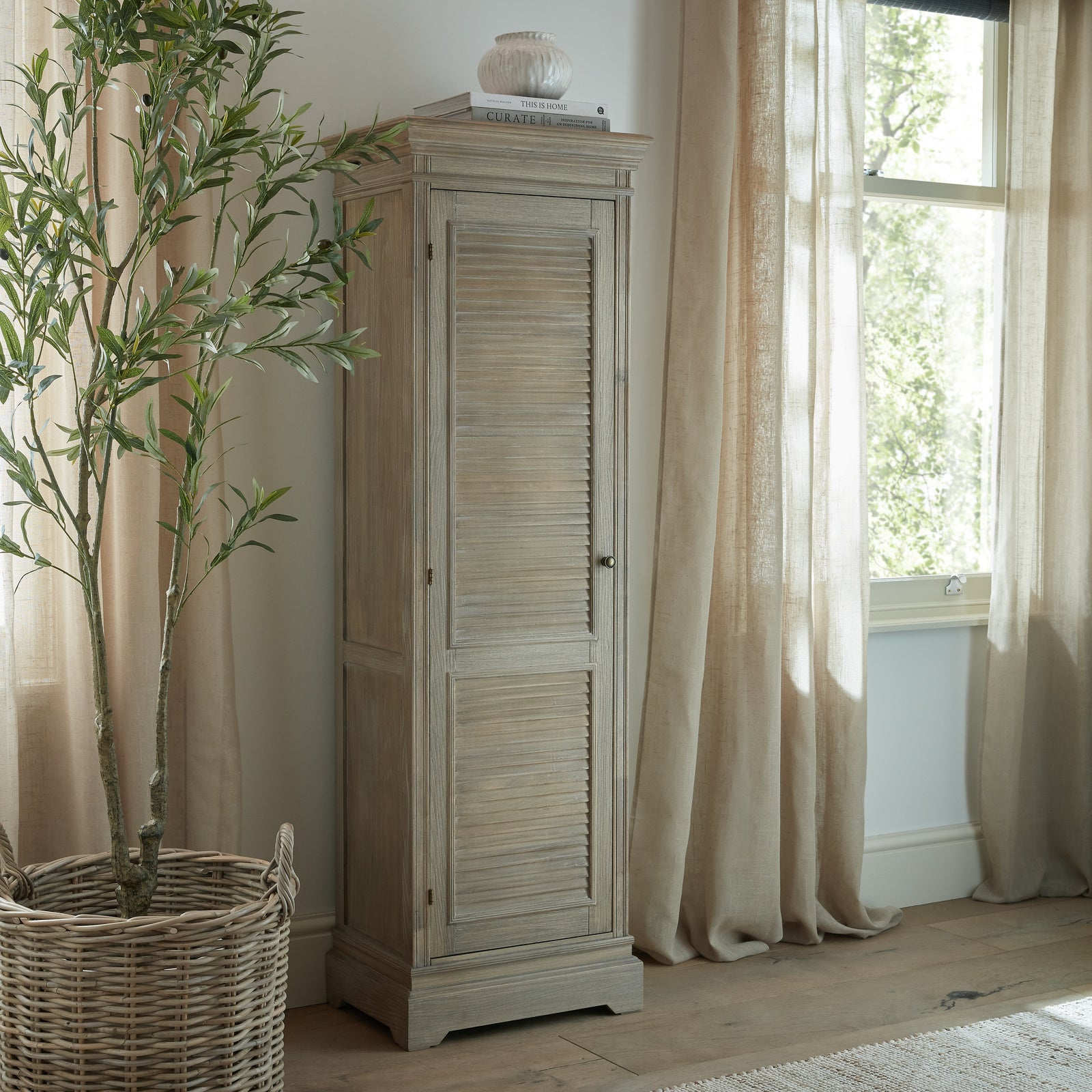 Laurent Tall Wood Storage Cabinet with 1 Door & 3 Shelves styled in a contemporary living room beside a window and indoor plant, showcasing its natural wood finish, tall profile and versatile storage for living rooms, hallways and bedrooms.