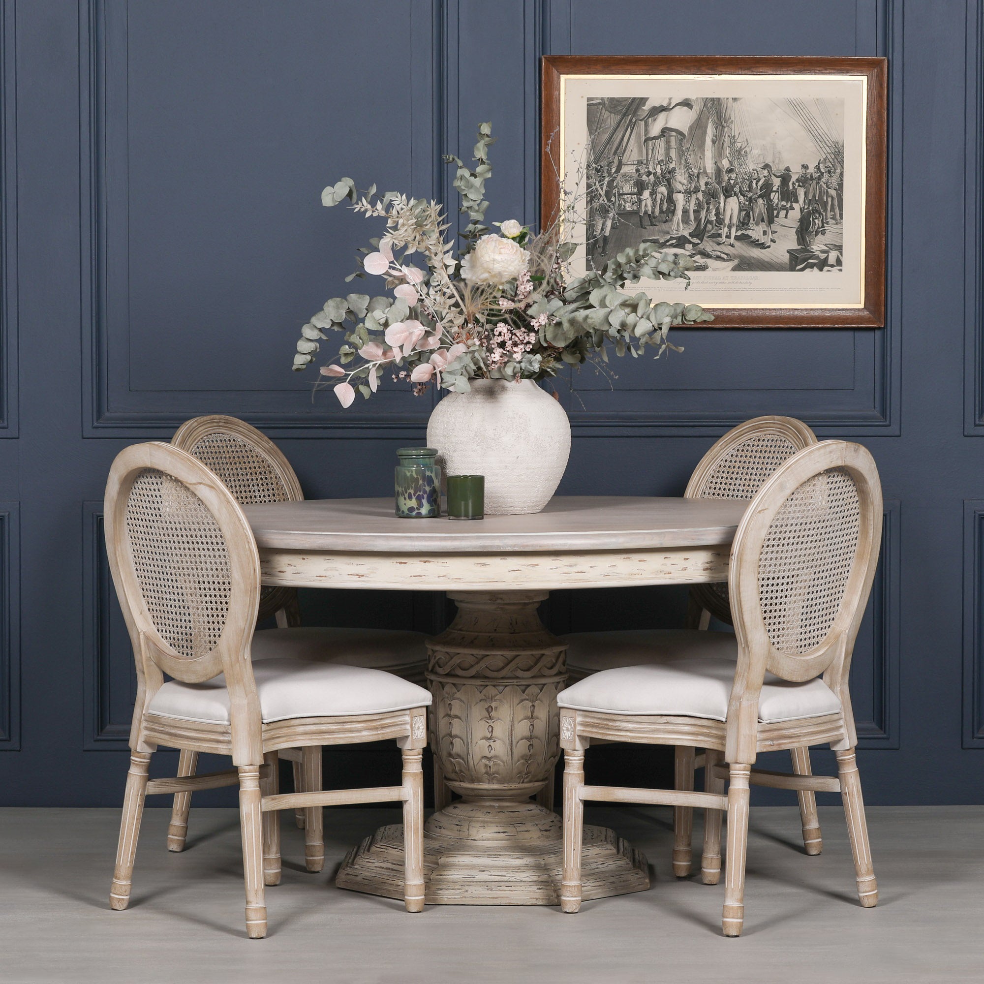 Léandre Round Pedestal Dining Table 145cm in distressed white cedar wood styled with four Louis upholstered dining chairs, decorative vase and accessories against a dark panelled wall, showcasing timeless handcrafted dining furniture.