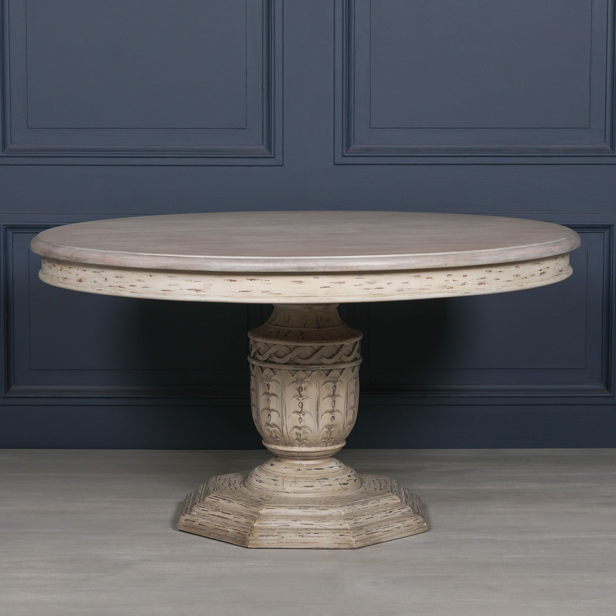 Front view of the Léandre Round Pedestal Dining Table 145cm featuring a distressed white cedar wood finish, spacious round tabletop and beautifully hand-carved pedestal base against a dark panelled interior.