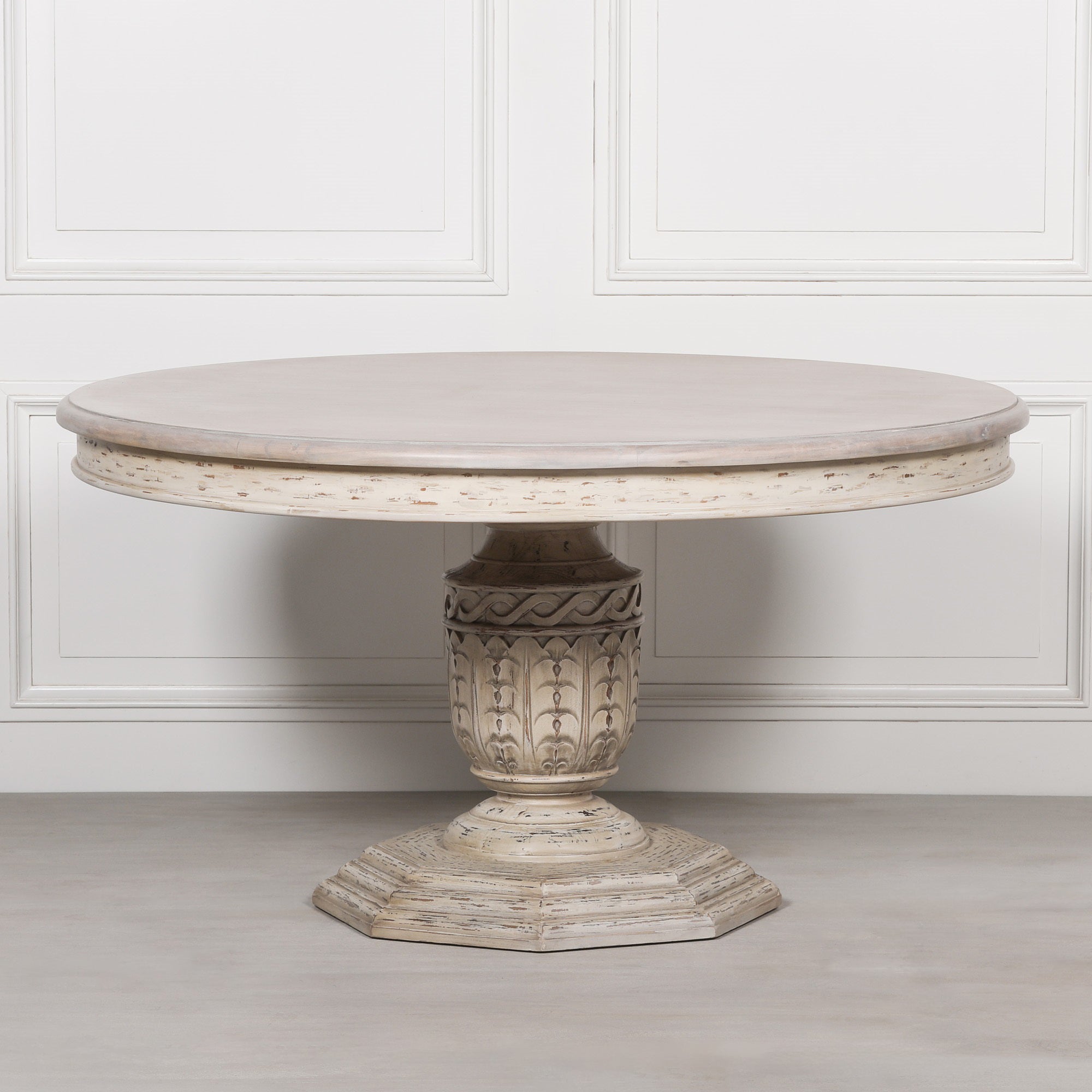 Léandre Round Pedestal Dining Table 145cm handcrafted from distressed white cedar wood with a generously sized round tabletop and intricately hand-carved pedestal base, shown against a white panelled wall.