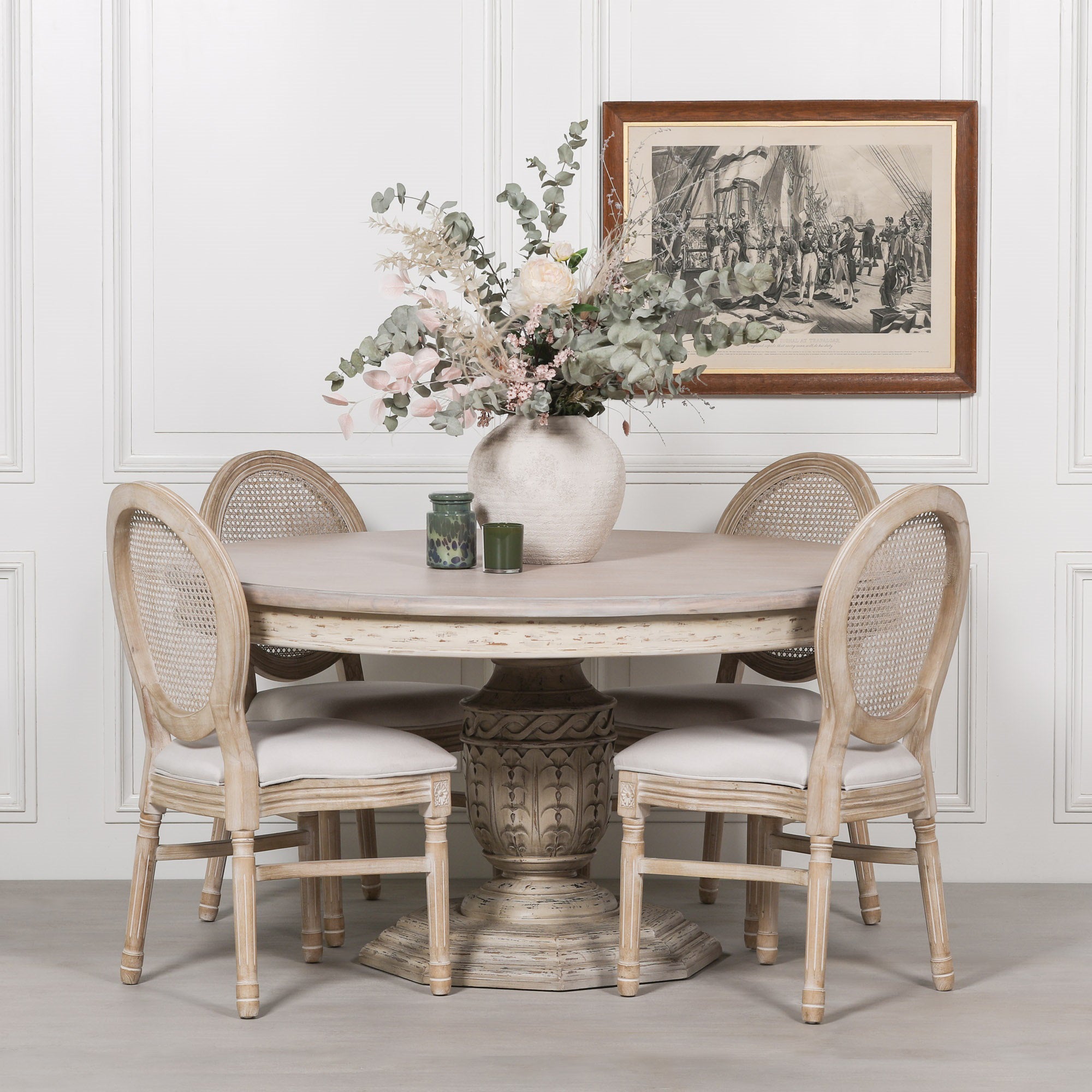 Léandre Round Pedestal Dining Table 145cm in distressed white cedar wood styled with four Louis upholstered dining chairs, featuring a hand-carved pedestal base, decorative floral arrangement and framed wall artwork in a classic dining room setting.