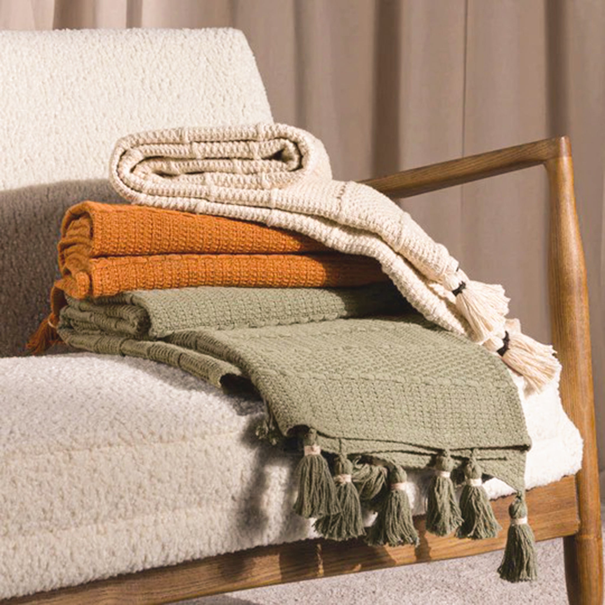 Léna Woven Cotton Tasselled Throw in Natural Beige styled with coordinating throws on an upholstered chair, showing its textured weave and decorative tassel detailing.