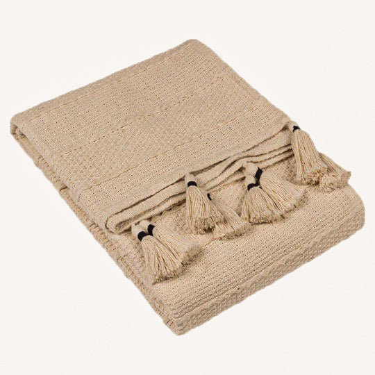 Folded Léna natural beige woven cotton throw with decorative tassel fringe, showcasing its neutral colour, textured weave and contrasting dark tassel ties.