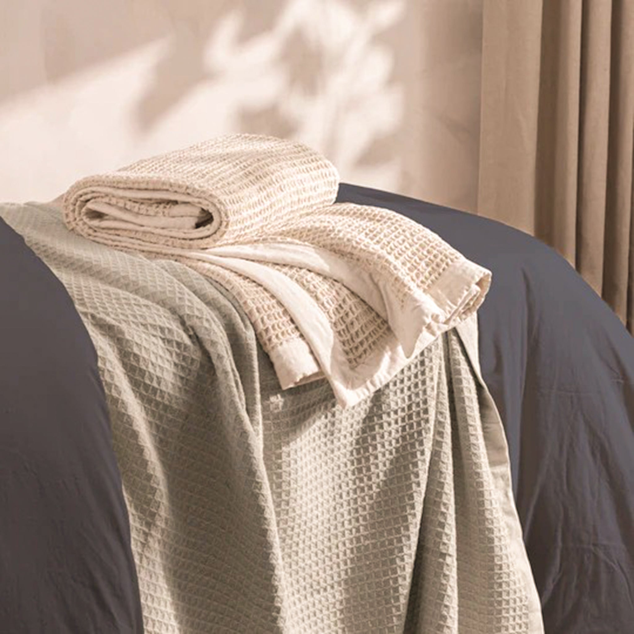 Léna Oversized Waffle Weave Cotton Throw in Stone & Natural layered across a bed, showing its generous size, textured cotton weave and soft neutral border in a warm bedroom setting.