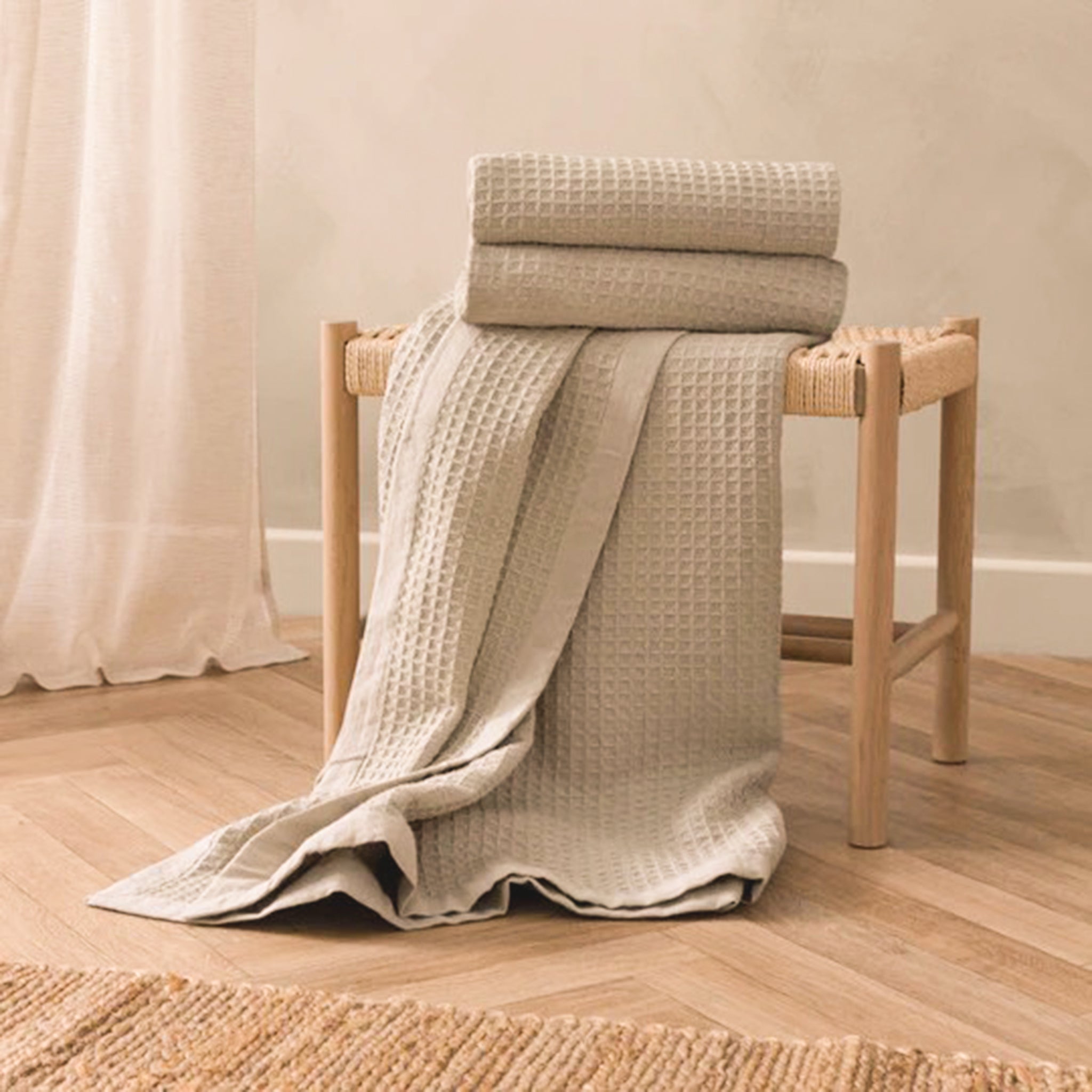 Léna Oversized Waffle Weave Cotton Throw in Stone & Natural draped over a wooden stool, showcasing its soft neutral colour, textured waffle weave and contrasting natural border.
