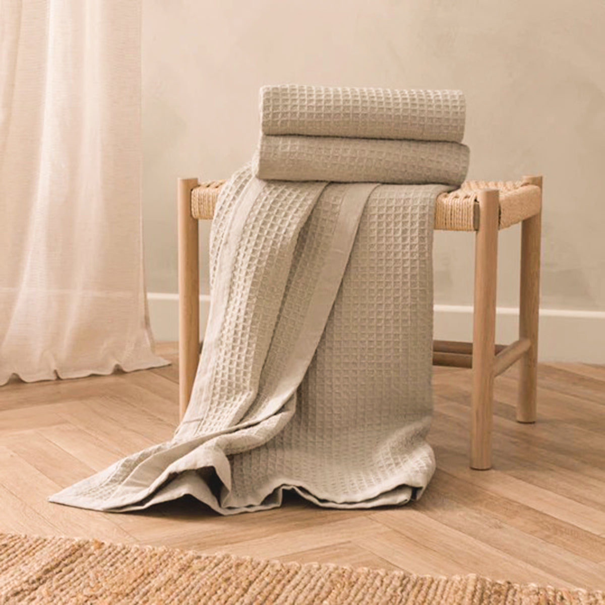 Léna Oversized Waffle Weave Cotton Throw in Stone & Natural draped over a wooden stool, showcasing its soft neutral colour, textured waffle weave and contrasting natural border.