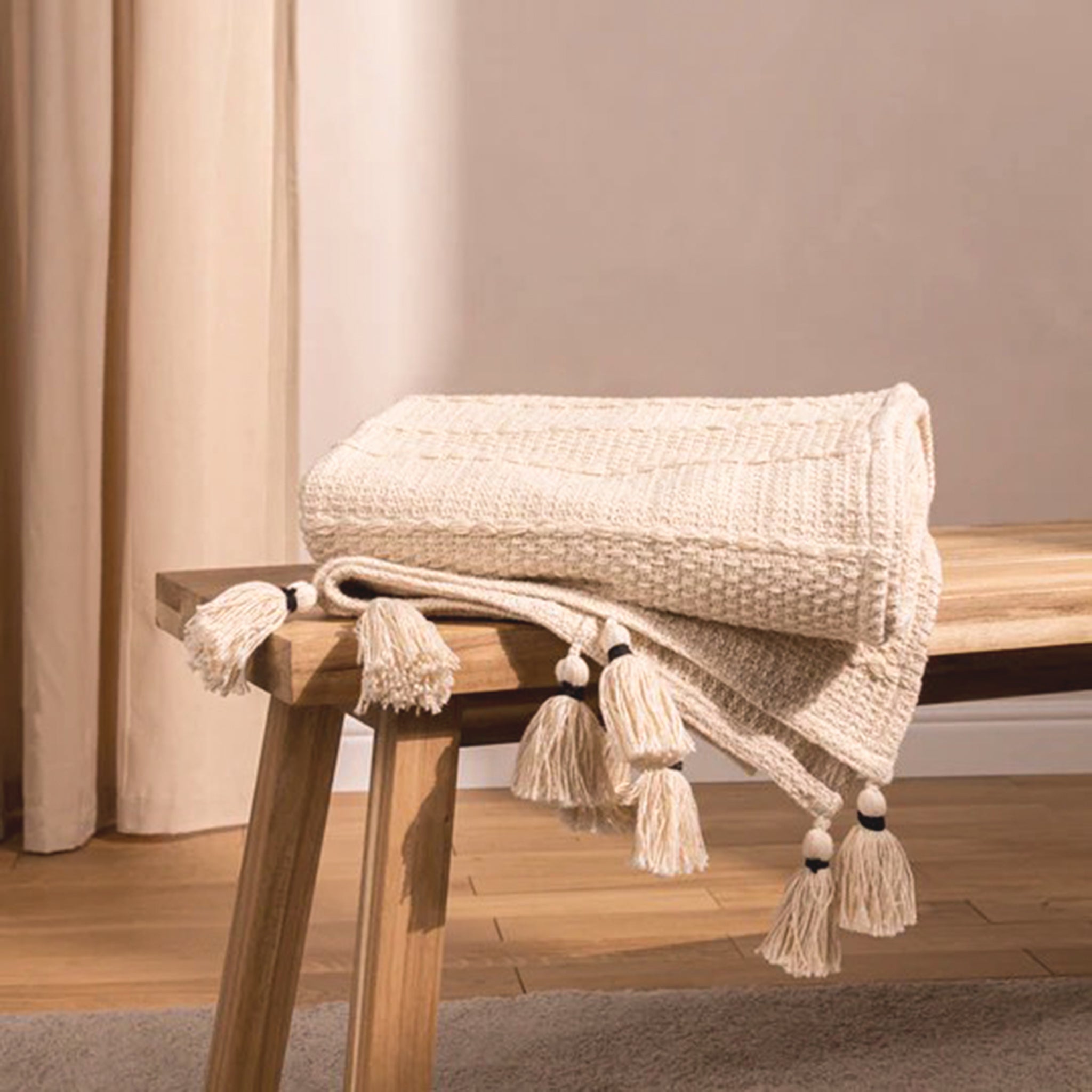 Léna Woven Cotton Tasselled Throw in Natural Beige folded over a wooden bench, showing its textured woven cotton design and decorative knotted tassel fringe.
