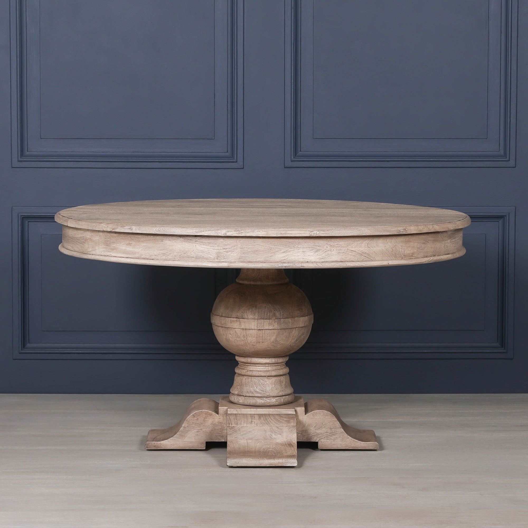 Front elevation of the Lucan round pedestal dining table crafted from solid rustic washed acacia wood, featuring a thick circular tabletop, turned ball pedestal and sculptural four-footed base against a dark navy panelled interior.