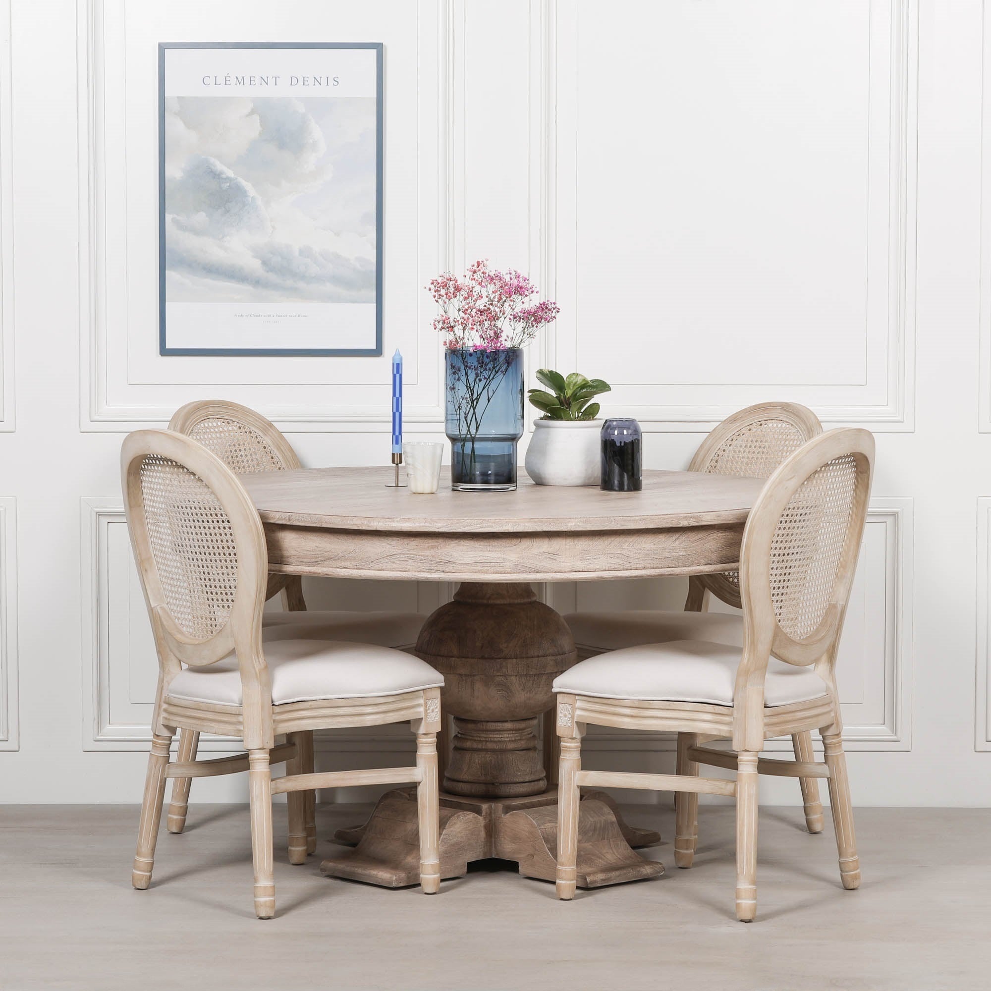 Round Lucan pedestal dining table handcrafted from rustic washed acacia wood, styled with four Louis upholstered dining chairs featuring natural rattan backs and linen seats. Displayed in an elegant dining room with white wall panelling, decorative floral arrangement and contemporary wall art.