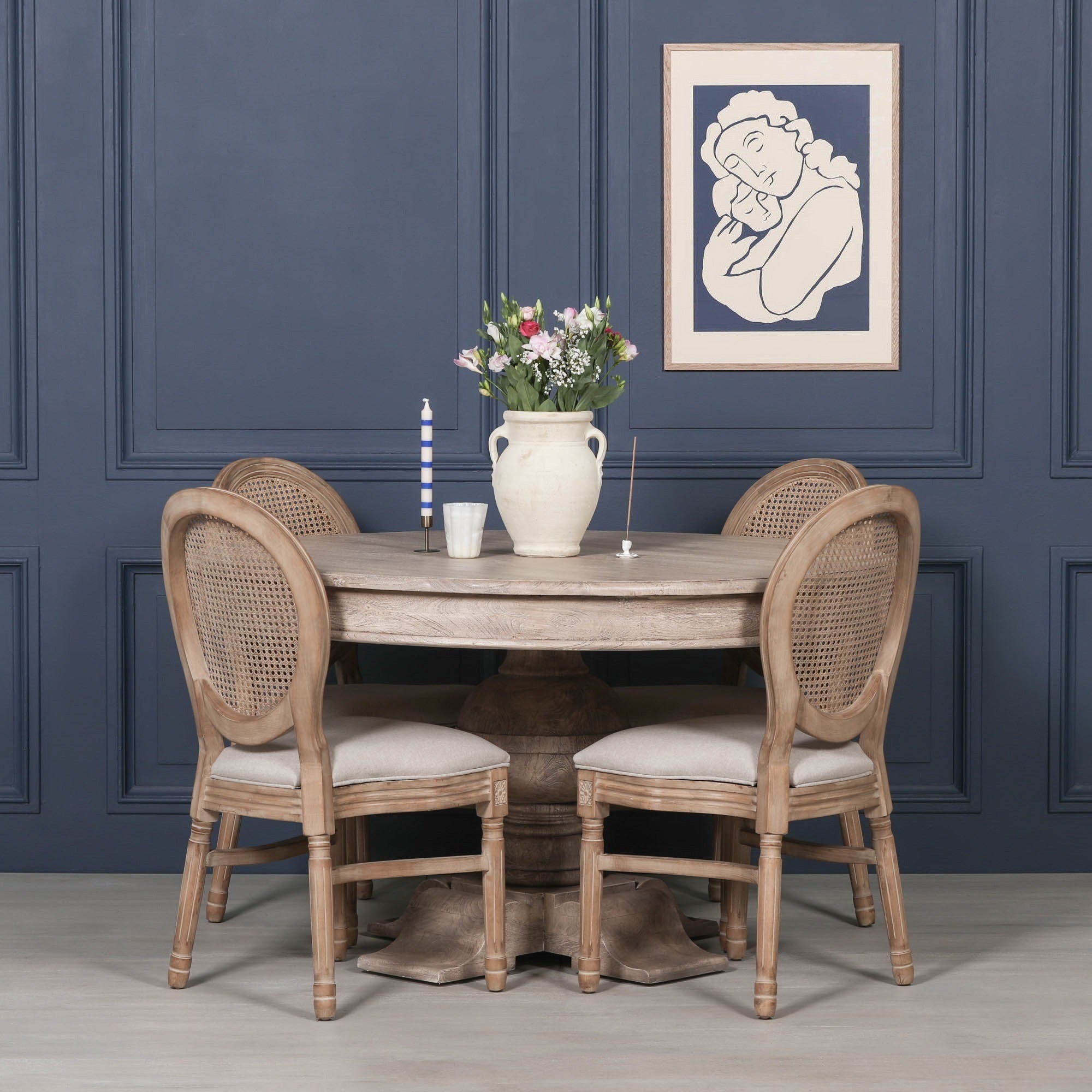 Lucan round pedestal dining table in rustic washed acacia wood styled with four Louis upholstered dining chairs in a sophisticated dining room featuring deep navy wall panelling, fresh floral arrangement and framed artwork.
