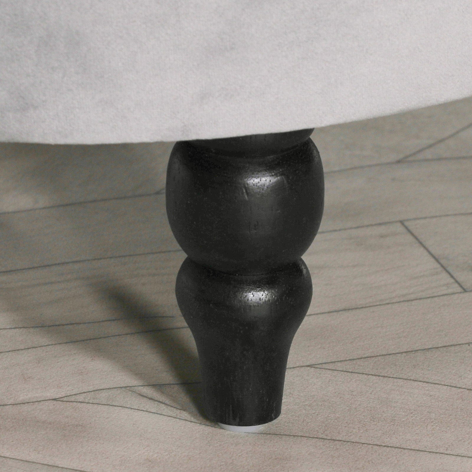 Close-up of Lucien bench black turned wood leg beneath grey velvet upholstery on a pale wood floor.