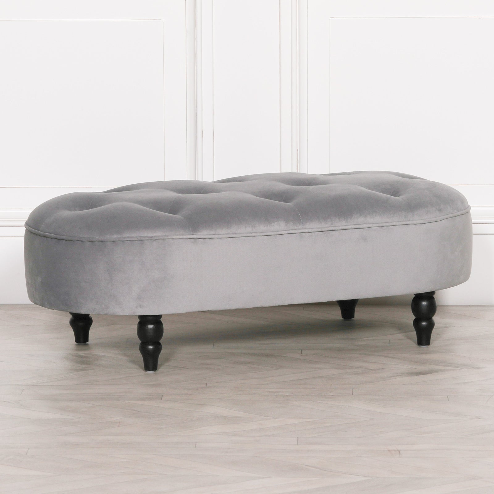 Grey velvet tufted bedroom bench with black turned legs against a white panelled wall and pale wood floor.