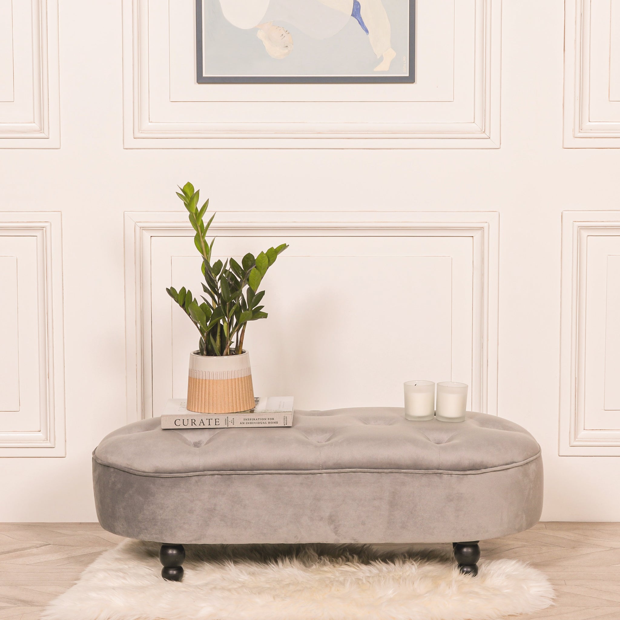 Lucien Grey Velvet Upholstered Bedroom Bench with Black Legs styled with decorative candles, books and a potted plant, showcasing its luxurious tufted velvet seat and elegant end of bed bench design for modern bedrooms and dressing rooms.