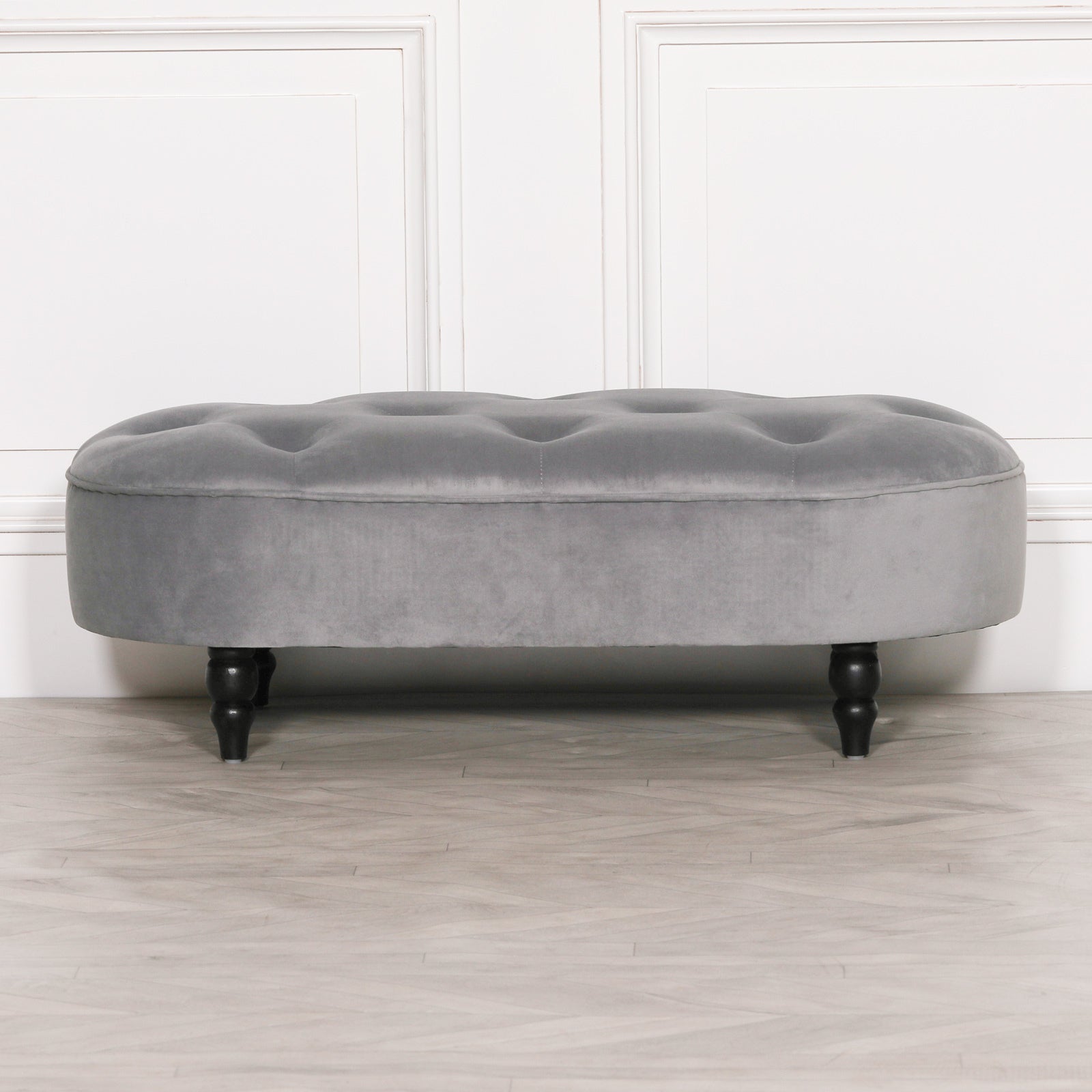 Grey velvet upholstered bedroom bench with deep tufting and black legs against a white panelled wall and wood floor.