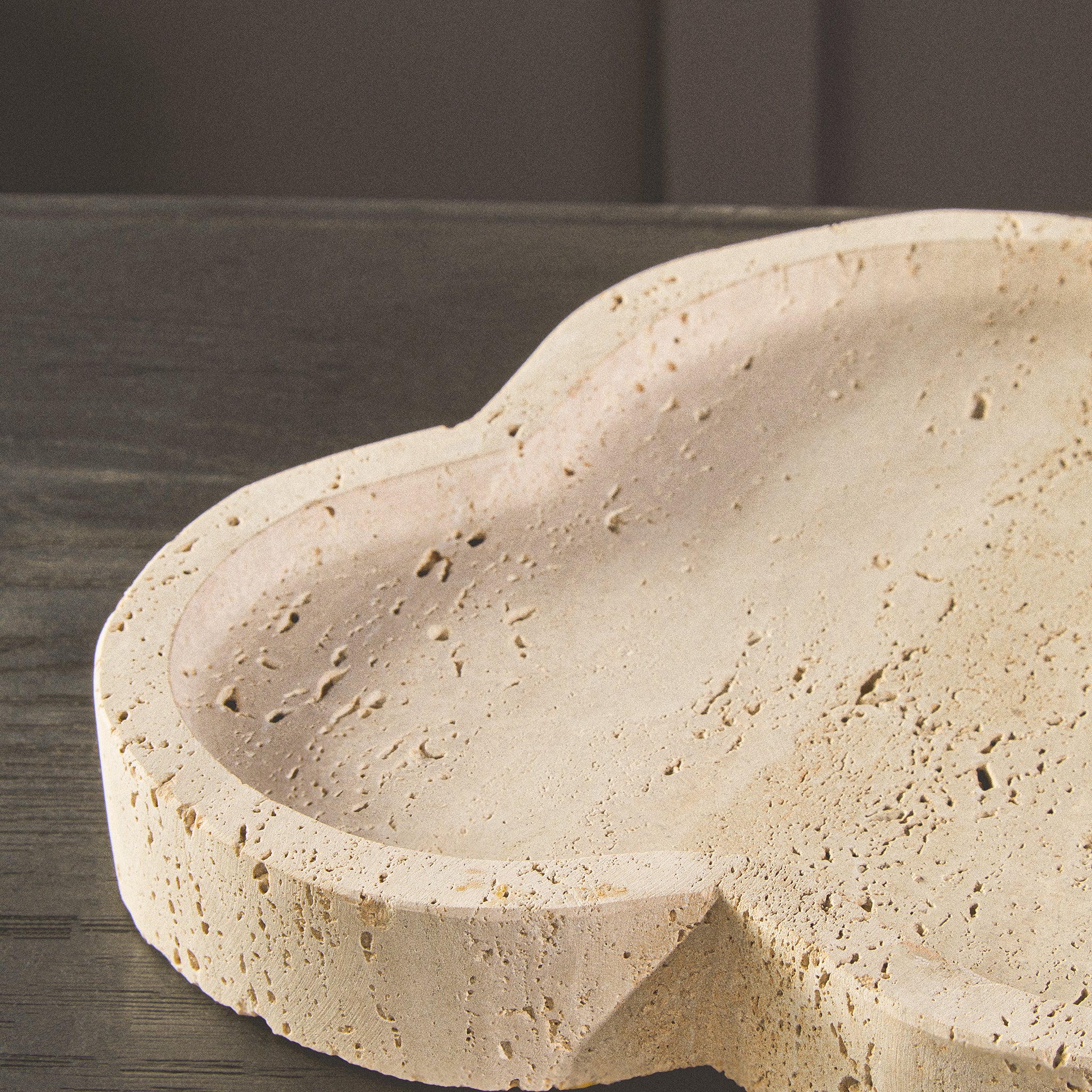 Close-up of the Noé Travertine Tray showing its irregular sculpted edge, raised rim and naturally porous beige stone texture on a dark wood surface.