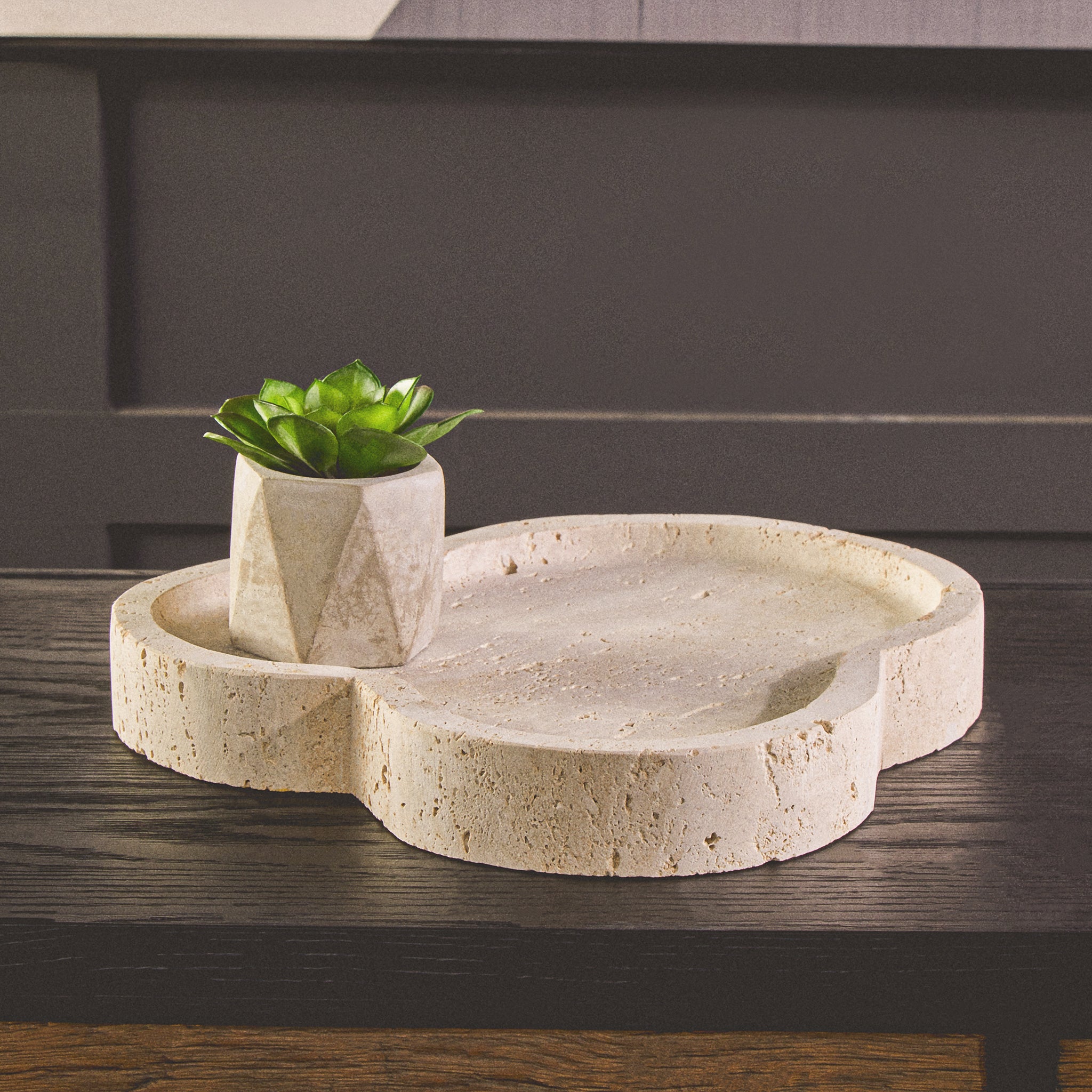 Noé irregular travertine decorative tray styled with a small potted plant on a dark wood surface, showing its sculptural organic shape and natural stone texture.