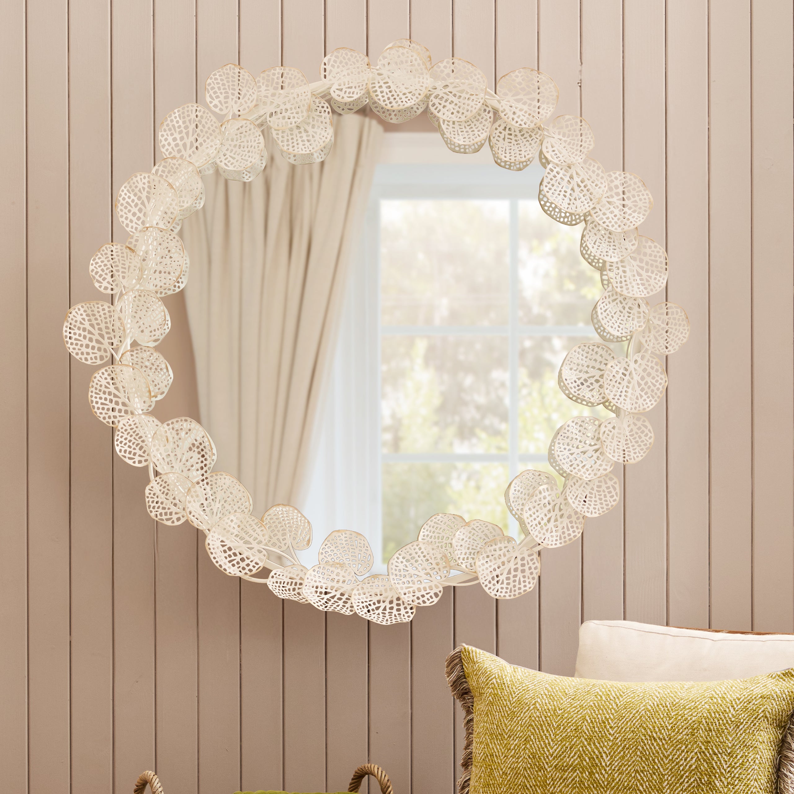 Odette 109cm Round Wall Mirror with antique white and gold floral frame displayed in a bright living room, reflecting natural light and adding an elegant decorative focal point above occasional furniture.