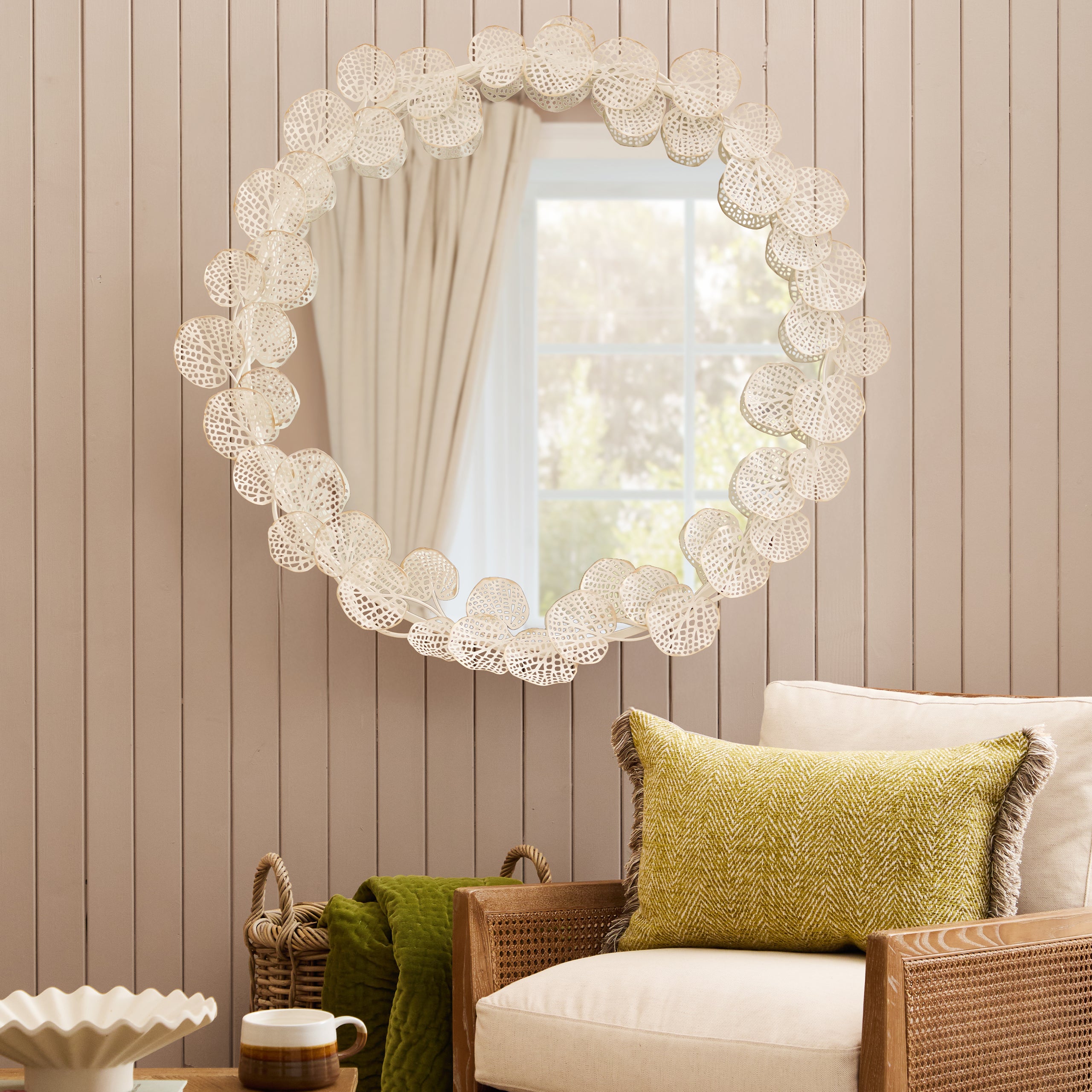 Odette 109cm Round Wall Mirror with an antique white and gold floral frame styled above a natural rattan accent chair, showcasing a luxury statement mirror for living rooms, bedrooms, hallways and elegant country-inspired interiors.