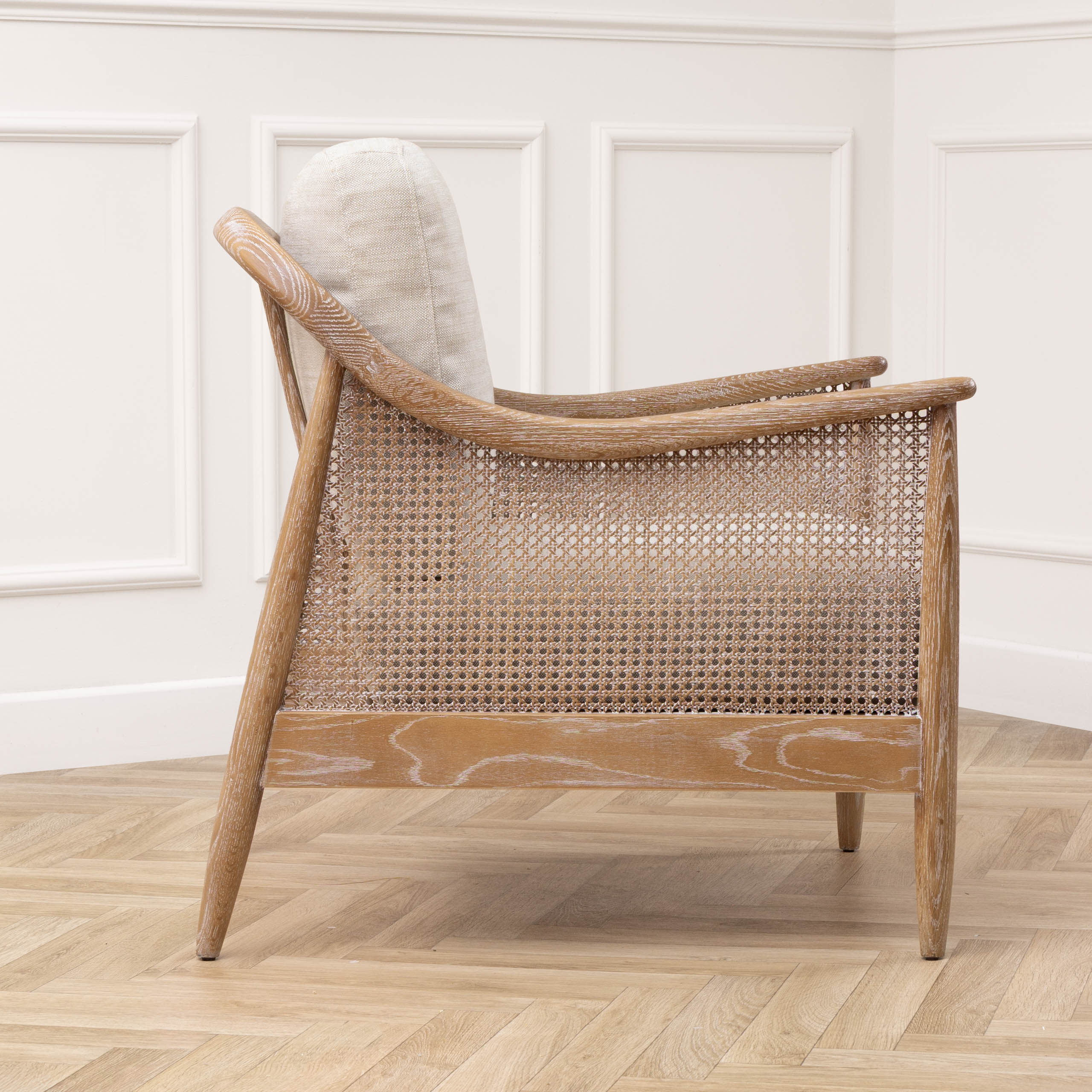 Side view of Remi linen 2-seater chair with woven rattan panel against white walls and herringbone floor.