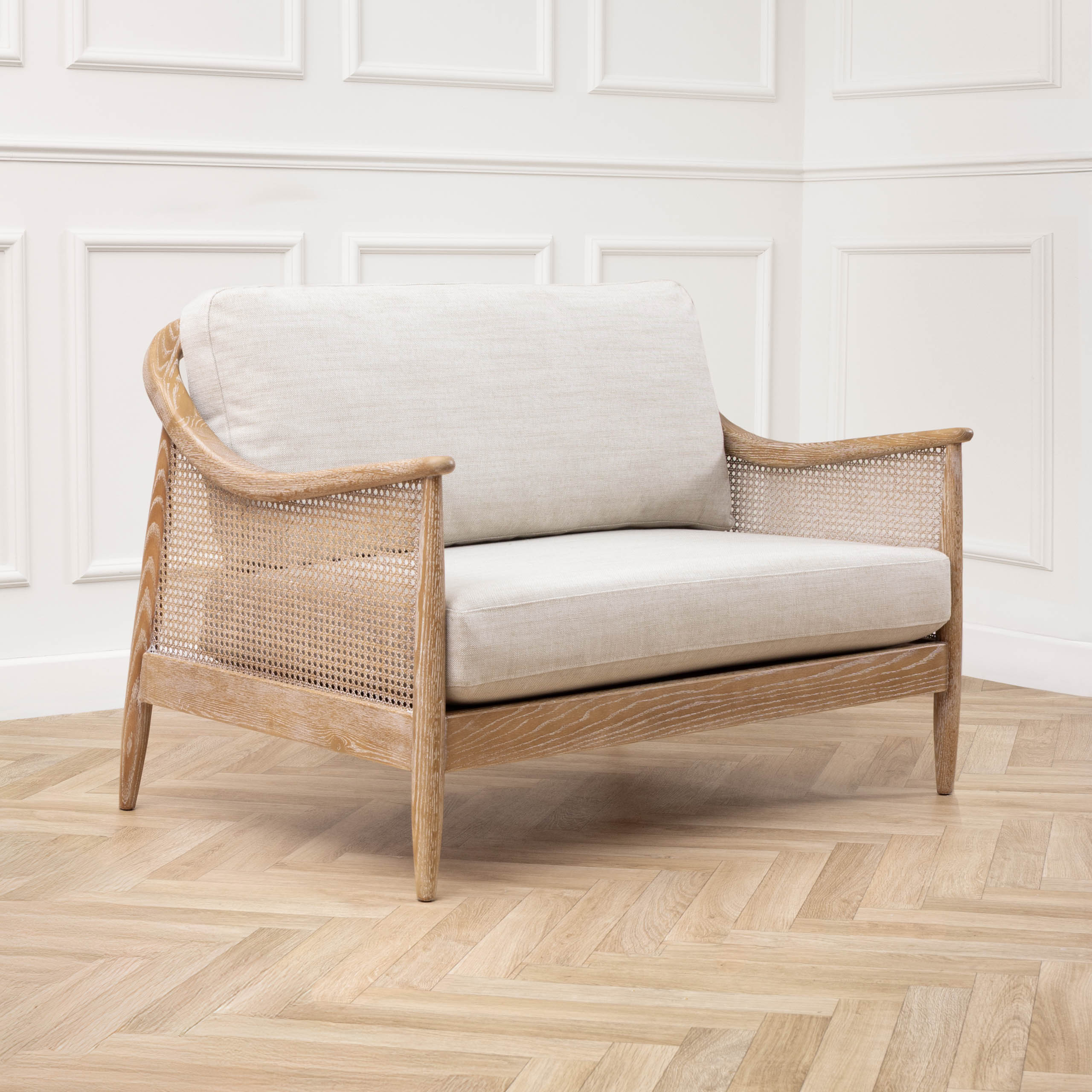 Remi rattan 2-seater chair with linen cushions against white panelled walls and herringbone wood flooring.