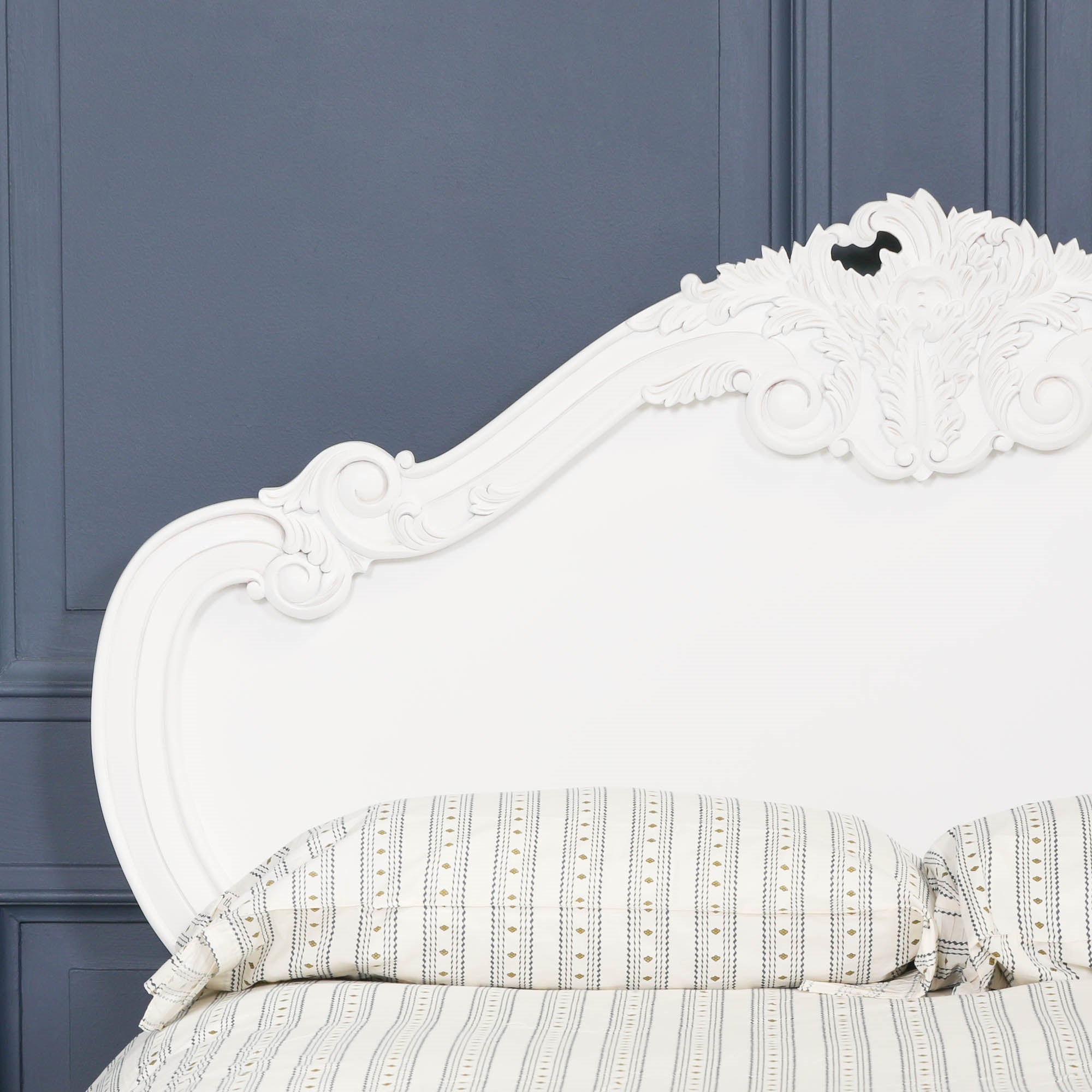 Close-up of the Rosalie French Rococo bed headboard with ornate hand-carved rose and cherub detailing in a light painted white finish against dark blue panelled walls.