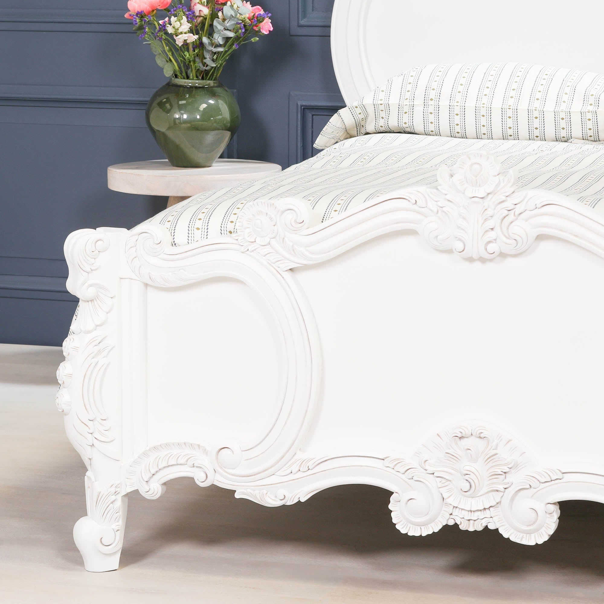 Close-up of the Rosalie French Rococo white double bed frame showcasing the hand-carved footboard with intricate rose and cherub detailing beside a bedside table with flowers.