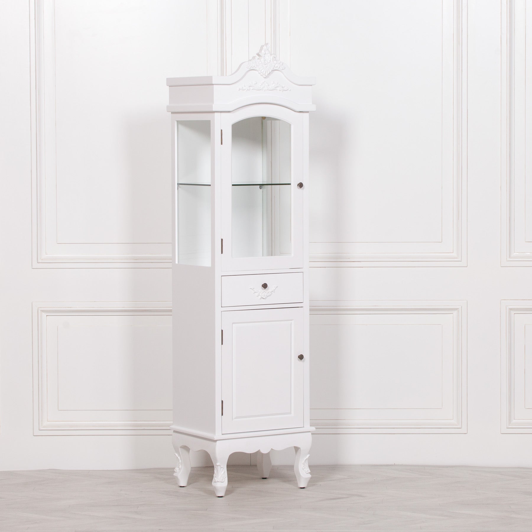 Angled lifestyle view of the Rosalie Tall French Glass Display Cabinet with Drawer in Antique White displayed in a bright French-inspired interior, highlighting its elegant proportions, glazed cabinet and decorative carved details.
