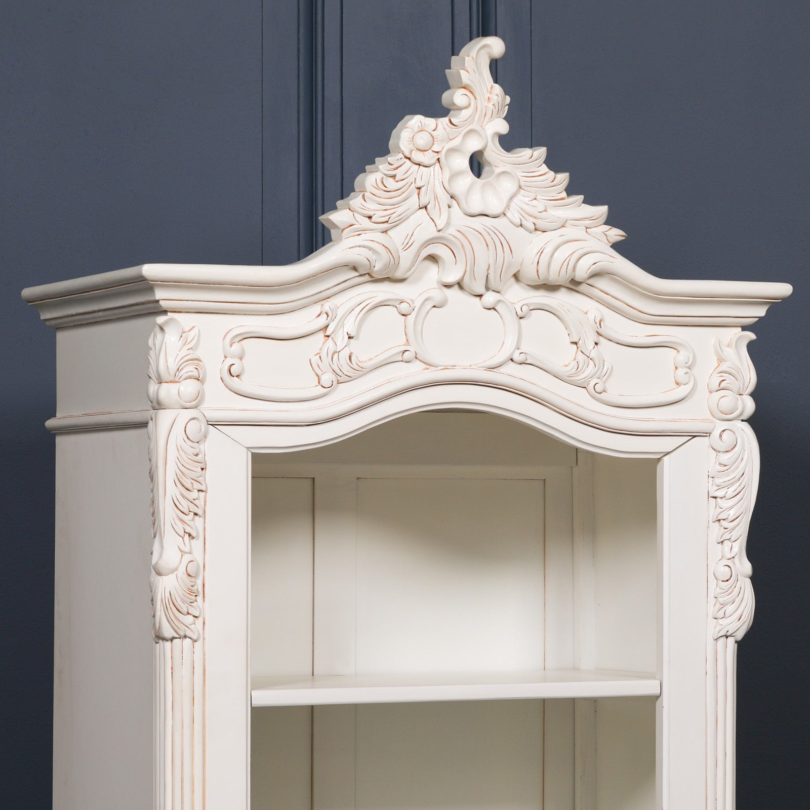 Close-up of the Rosalie Tall French Open Bookcase showcasing the elaborate hand-carved Rococo crown, floral crest, scrolling ornamentation and Antique White mahogany wood craftsmanship inspired by classic French furniture.