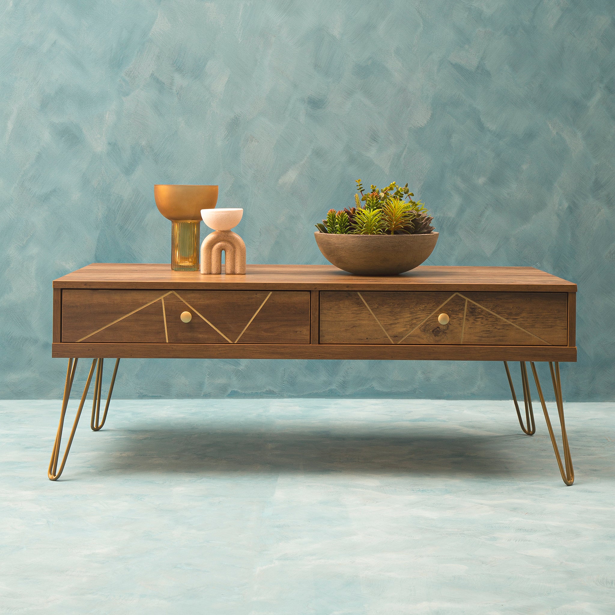 Soline wooden coffee table with two geometric-front drawers and gold hairpin legs styled with decorative objects against a textured blue interior wall.
