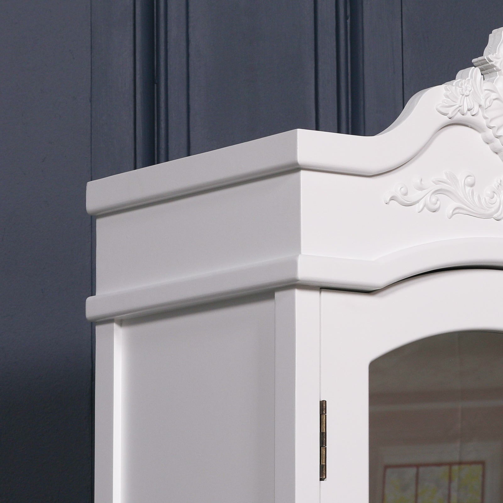 Side close-up of Vivienne white armoire carved crown and mirror frame against a dark navy panelled wall.