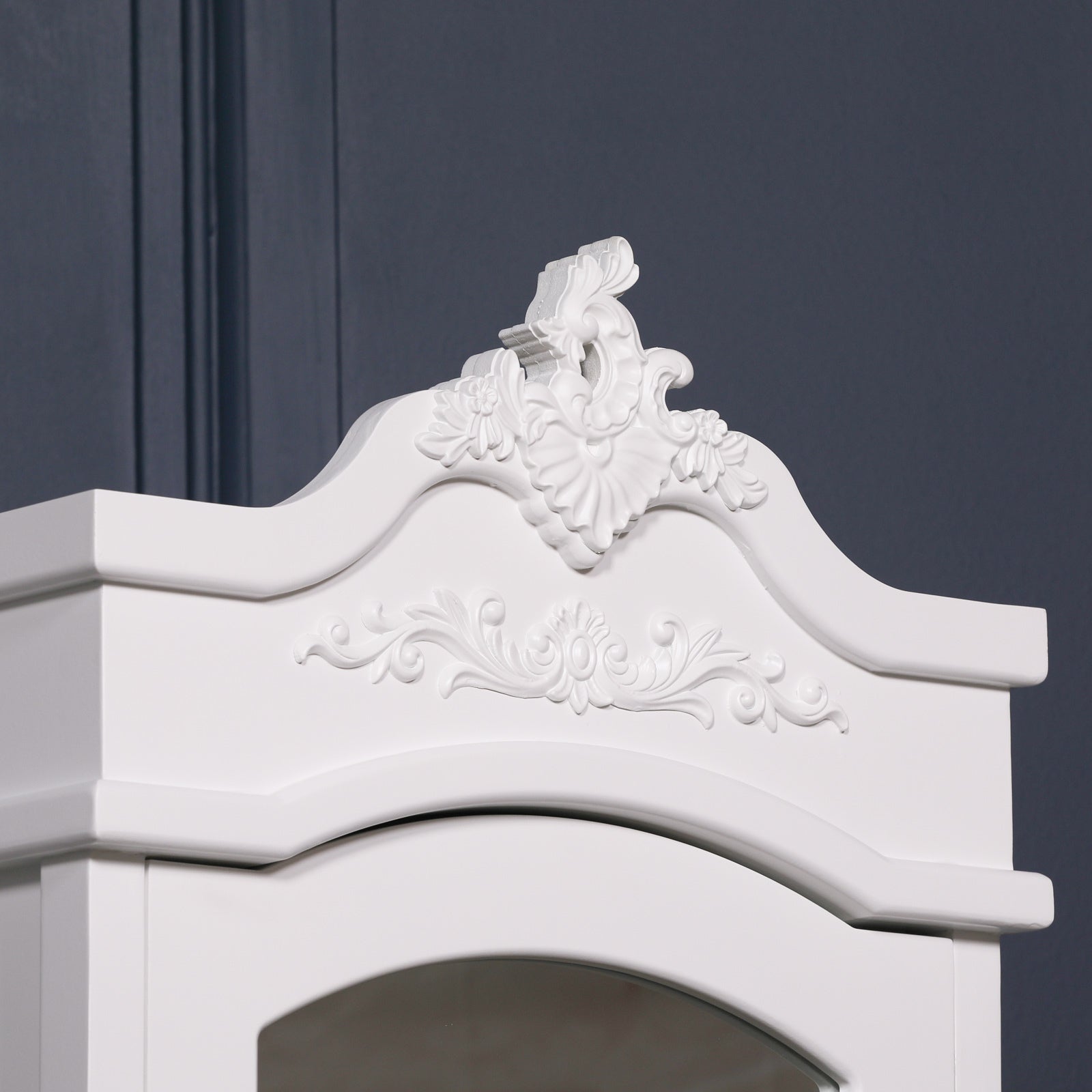 Vivienne white mirrored armoire ornate carved top detail against a dark navy panelled wall.
